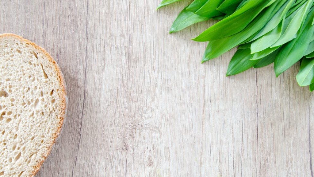 A slice of bread and fresh wild garlic leaves on a rustic wooden table.