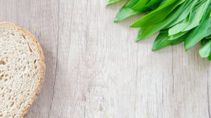 A slice of bread and fresh wild garlic leaves on a rustic wooden table.